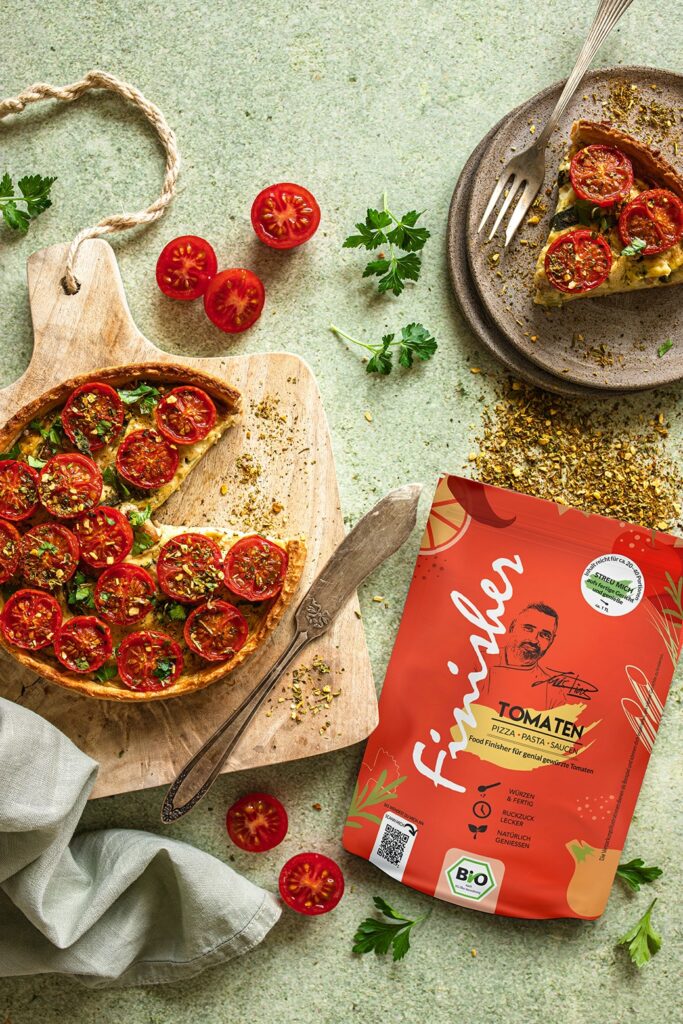 Fladenbrot mit Ricotta, Artischocken und Tomaten auf einem Holzbrett, daneben ein Teller mit einem Stück davon sowie die rote Verpackung des Bio Antipasti Food Finishers.