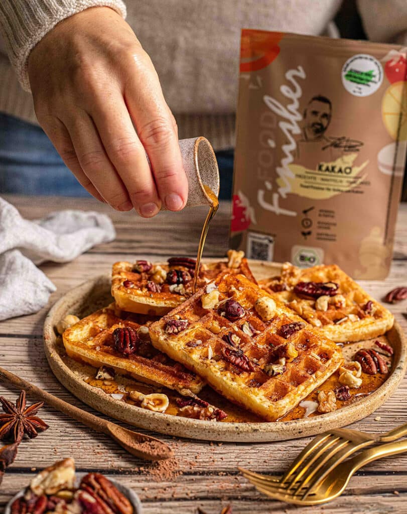 Eine Hand gießt Sirup über knusprige Kürbiswaffeln, die mit Nüssen garniert und mit BIO KAKAO FOOD FINISHER bestäubt sind. Die Waffeln liegen auf einem rustikalen Teller, der auf einem Holztisch angerichtet ist. Im Hintergrund ist die Packung des BIO KAKAO FOOD FINISHER sichtbar.