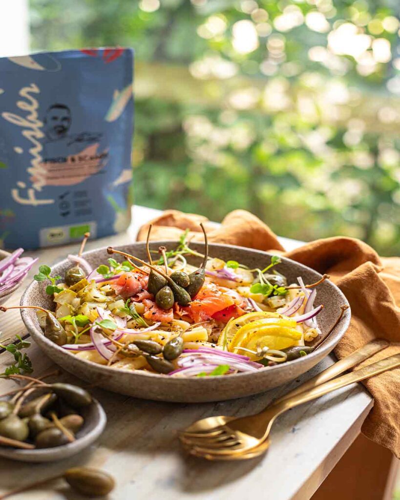 Nudelsalat mit gelber Paprika, roten Zwiebeln, Kapernäpfeln und Hähnchenstreifen in einer rustikalen Schale. Im Hintergrund ein natürlicher Garten und die Verpackung des Bio Fisch & Scampi Food Finishers.