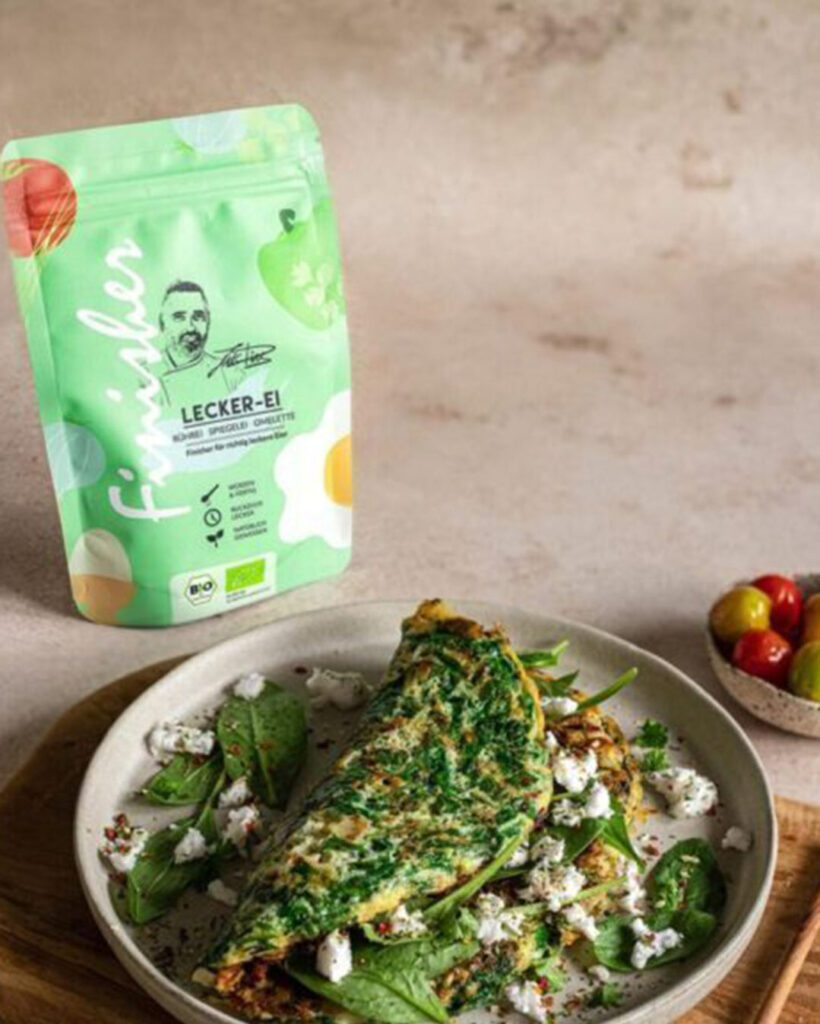 Ein goldbraunes Omelette mit Spinat und Feta auf einem Teller, daneben die grüne Verpackung des Bio Lecker-Ei Food Finishers mit der Illustration von Koch Luis Dias. Umgeben von frischen Zutaten und Gewürzen.